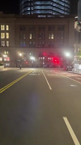 The video shows an urban street scene at night. A wide street with painted lane markings and "ONLY" printed in the lanes faces a brick building with lighted windows and a taller modern skyscraper behind it. A red traffic light is visible. There is a bus stop on the right side of the street, with a person standing within it. A car is also parked on the right side of the street. Streetlights illuminate the scene.