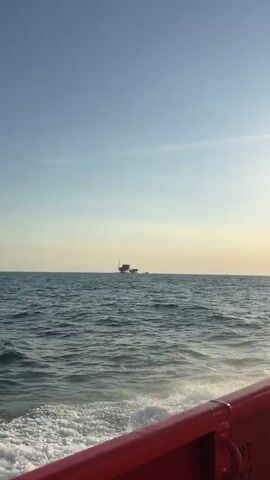 The video shows a view from a boat moving through the ocean. The water is choppy with white foam trailing behind the boat. In the distance, there is a large oil rig or offshore platform. To the right of the platform, a small boat is moving across the water. The sky is clear with a gradient from blue to a lighter hue near the horizon, indicating it might be early morning or late afternoon. The perspective suggests the viewer is on a vessel that is moving towards or parallel to the oil rig.