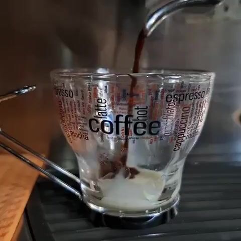 Dark liquid is poured into a clear glass from a metal faucet. The glass has white liquid at the bottom and is printed with coffee-related words in various fonts and colors. The glass is sitting on a metal grate on a beige surface.