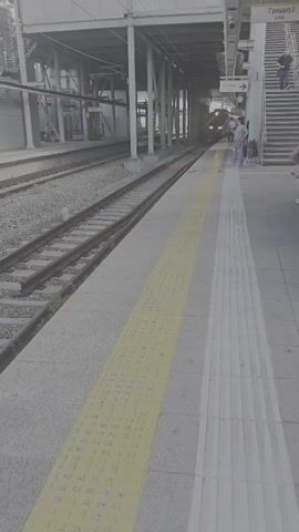 The video shows a train arriving at a station. The train is white and purple. There are several people standing on the platform, waiting for the train. One person is walking up a set of stairs. The platform has tactile paving to indicate the edge. The station appears to be elevated.
