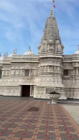 The video begins with a close-up shot of a white, intricately carved temple with multiple domes and a tall spire topped with a flag. The foreground is paved with red and tan interlocking tiles. A young woman in a green shirt and blue jeans stands on the right side of the frame, holding up her phone to take a picture of the temple.

The camera then pans to the left, revealing more of the temple's exterior and the surrounding grounds. The temple architecture is detailed with carvings of figures and patterns. A walkway leads to an entrance with a dark wooden door. To the left of the entrance, a white urn filled with plants is visible.

As the camera continues to pan, the view expands to show a large, manicured lawn with several fountains. The fountains have water shooting upwards in arcs. In the middle of the lawn, a person in an orange shirt is operating a lawnmower. Several other people are walking in the distance near the temple.

The video concludes by showing a wider view of the temple grounds, including lush green lawns, trees in the background, and decorative archways. The sky is blue with scattered white clouds. The overall scene depicts a serene and well-maintained religious complex.