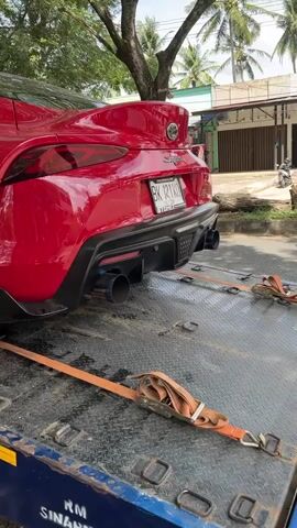 A red 2020s Toyota Supra is strapped to a tow truck. The car has aftermarket wheels and exhaust. The tow truck is parked on the side of a tree-lined road. There is a building visible in the background.
