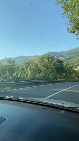 This is a first-person perspective video taken from inside a car driving on a road in Podličak, Montenegro. The scene is a scenic route with a clear blue sky and bright sunshine, indicating a pleasant day. The road is paved with white lane markings, and a metal guardrail lines the side of the road. Lush green trees and foliage are abundant, with a mountainous landscape dotted with buildings visible in the background. The time of day appears to be late afternoon, given the angle of the sunlight casting shadows.

The activity captured is a car journey through a picturesque, hilly area. Several other vehicles are visible on the road, including a red car driving ahead of the viewpoint car and a few other cars further down the road. The buildings in the background are a mix of residential and possibly commercial structures, nestled into the hillsides, suggesting a town or village. The overall mood is one of travel and enjoying a scenic drive. No specific individuals are clearly discernible within the vehicle. There is no visible text in the video.