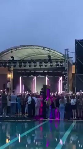 A crowd of people gather at dusk in front of an outdoor stage where a band is performing. The crowd is relatively dense, and many individuals are holding up their phones, presumably to take photos or videos of the performance. In front of the crowd is a swimming pool with turquoise water. The stage is covered and brightly lit, with geometric light installations. There are at least four performers on stage, visible through the lights and scaffolding. The overall atmosphere suggests a concert or outdoor event.