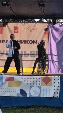 A man in a black and white shirt and black pants sings into a microphone on a stage. To his right, a woman in a black, sheer dress with lace detailing and black stockings dances. They are performing in front of a backdrop that has text and logos. A purple curtain is to the right of the backdrop. The stage has a banner with colorful geometric shapes along the front. The scene appears to be outdoors.