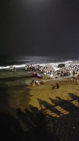 The video shows a crowded beach at night. The water is visible on the left, with waves and a dark sky above. A large number of people are gathered on the sand, some near the water's edge and others further inland. There are tents, tables, and what appear to be some small structures on the beach. Shadows are cast on the sand in the foreground, suggesting artificial lighting. Several palm trees are visible on the right side of the frame. The overall scene depicts a busy nighttime gathering on a beach.
