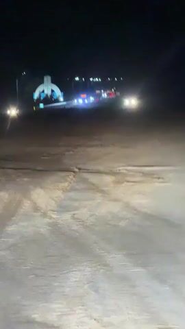 The video shows a nighttime scene with a road in the foreground and a large illuminated structure in the distance. The road appears to be unpaved or covered in sand or dirt, with tire tracks visible. Several vehicles are on the road, their headlights creating beams in the darkness. The illuminated structure in the background is a large, arch-like monument with blue and white lights. There are also other smaller lights and structures visible behind the monument, possibly buildings or streetlights. The sky is dark and clear, indicating it is nighttime. The overall impression is one of a somewhat desolate but lit-up area at night, possibly a roadside attraction or entrance to a town or facility.
