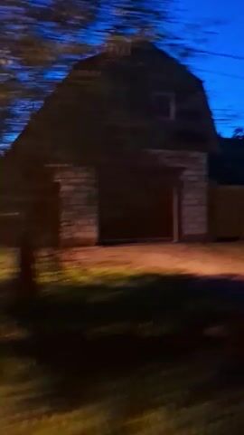 The video opens on a dark evening with a shot of a two-story building with a gambrel roof. The building appears to be a garage with a large, dark-colored door. Two square windows are visible on the upper level. The camera then pans to the right, revealing a wooden fence. The scene is illuminated by artificial lights casting an orange glow on the ground. The camera continues to pan right, showing trees and foliage against a dark blue sky. The lighting shifts, with parts of the scene becoming illuminated with green light, possibly from a different light source. The video ends with a view of a yellow wall with an arched opening, partially obscured by trees. The overall impression is that of a residential backyard or property at dusk or night.