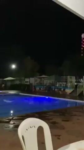 The video shows a swimming pool area at night. The pool is illuminated with blue lights. There is a lifeguard stand next to the pool, as well as a yellow bus nearby.  The video is taken from a position underneath a covered patio area.  A white plastic chair is partially visible in the foreground.  There is music playing in the background.