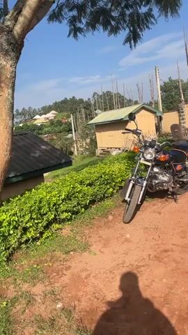 A motorcycle is parked on a dirt path in a rural setting. The motorcycle is black with orange and blue accents. In the background, there are several buildings, including a small yellow structure with a green roof and a larger building with a dark roof. A lush green hedge runs along the left side of the path. Beyond the buildings, rolling hills are covered with trees and some houses. The sky is blue with some white clouds, suggesting it is daytime and likely fair weather. A shadow of a person is visible on the ground in the foreground, indicating someone is taking the photo. The overall scene suggests a peaceful, perhaps slightly remote, rural environment in Gembu, Nigeria.