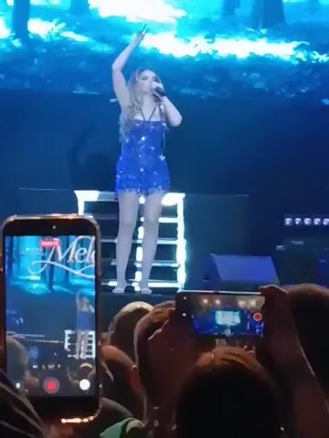 A concert attendee films a performance of a singer in a blue dress on a stage with a digital backdrop of trees. The singer gestures with her arms outstretched towards the audience who respond with cheers and enthusiasm. The camera then moves, focusing on the faces of people watching the performance.