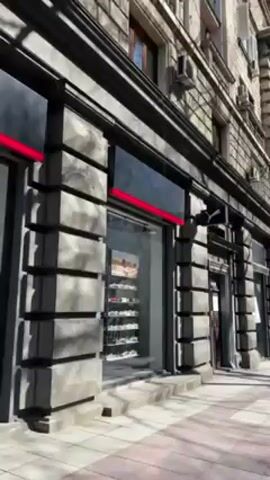 The camera pans to the right, showing a city street on a sunny day. The sidewalk is paved with gray square tiles. On the left is a long, gray stone building with storefronts. One of the businesses is a Puma store with shoes displayed in the window. Trees line the street. A black iron fence runs along the edge of the sidewalk, separating it from the street. Cars are parked along the street, and some are driving by. A person walks in the distance along the sidewalk. The sky is blue and cloudless.