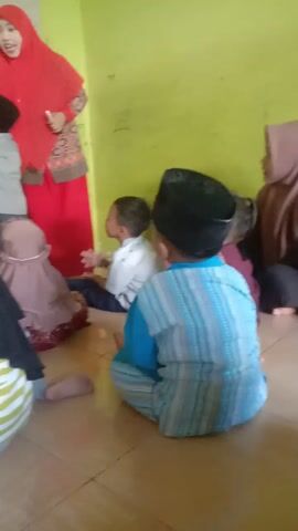 The video shows a group of young children and at least one adult in a room. The room has a light green wall and a tiled floor. Several children, dressed in what appear to be Islamic attire including hijabs and skullcaps, are on the floor in various prayer positions. One child in the foreground, wearing a black hijab and a striped shirt, is looking towards the camera with a smile. Another child in a red hijab is standing and appears to be instructing or interacting with the children. The children are positioned on a light-colored mat or carpet. The overall scene appears to be a religious education or prayer session for young children.