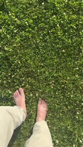 The video shows a first-person perspective of a person walking barefoot on a grassy area. The grass is green and appears to be a lawn with small white flowers scattered throughout. The person's feet are visible, with the left foot stepping forward first, followed by the right. The person is wearing light-colored pants. The sunlight casts shadows on the grass, indicating it is daytime. The camera is held relatively still, following the movement of the person's feet as they walk forward.