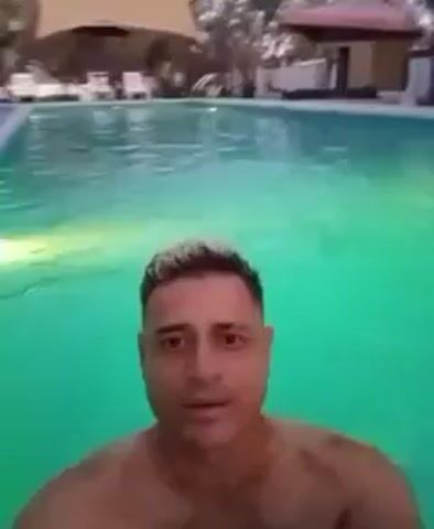 A man films himself in a swimming pool. The water is illuminated with colorful lights, changing from green to blue to pink. The man has short, light-colored hair and is looking directly at the camera. Behind him, the edge of the pool and some buildings or structures are visible. The overall setting appears to be an outdoor area at night or dusk.