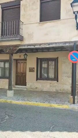 The video shows a street scene with traditional European architecture. The buildings have stone foundations and upper levels with exposed wooden beams. There are windows, doors, and a balcony visible. A street lamp and a no parking sign are present. Paved sidewalks and a street with a yellow line are also seen. Several cars are parked along the street, including a silver minivan and a red sedan. In the background, more buildings and a clear blue sky are visible. The scene appears to be in a quiet town or village.