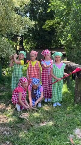 A woman in a white patterned shirt and beige shorts directs a group of six young girls dressed in matching checkered overalls and headscarves. The girls are in an outdoor setting with lush green trees and grass. Some of the girls are holding props, one has a bundle of sticks, another a red shovel. Two girls are crouching down in the foreground examining something on the ground. The woman appears to be giving instructions or demonstrating something to the girls. The overall atmosphere is one of organized play or a rehearsal.