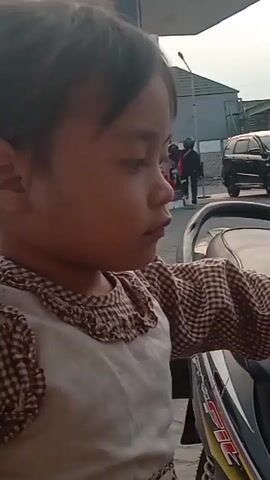 A young child with dark hair tied in a ponytail is shown wearing a brown and white checkered dress with a ruffled neckline. The child is sitting on a motorcycle. The background shows a street scene with a building, a car, and a motorcycle with a rider. The child looks directly at the camera and then turns their head to the side, opening their mouth. The lighting suggests it is daytime.