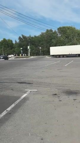 A large white truck is parked in a parking lot. The truck has a long, rectangular trailer attached to it. The parking lot is paved and has white lines marking the parking spaces. In the background, there are trees and a building. The sky is blue with some clouds.