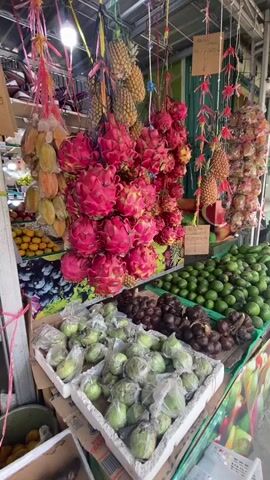 The video shows a vibrant fruit stall. In the foreground, there are white styrofoam containers filled with green guavas, some of which are wrapped in plastic bags. To the right of the guavas, there are rows of small, dark brown fruits, possibly salak or snake fruit. Further to the right, a large display of green limes or similar citrus fruits is visible. Hanging above the stall are bunches of bright pink dragon fruits, pineapples, and yellow star fruits. In the background, more fruits are displayed, including oranges and what appear to be mangoes. The stall is covered by a metal roof structure, and the overall environment suggests an outdoor market or a covered market setting. There are no people clearly visible in the frame.