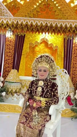 A woman adorned in traditional wedding attire, featuring an ornate gold headdress, a deep red, gold-embroidered jacket and skirt, and significant gold jewelry, is seated in a white ornate chair. She is positioned in front of a lavish backdrop characterized by gold fabric, intricate gold trim, and burgundy drapes. The woman smiles and waves at the camera in a series of clips. The scene suggests a formal cultural celebration, likely a wedding.