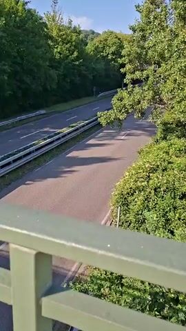 The video captures a view from a bridge overlooking a two-lane road with guardrails on both sides. Dense green trees and foliage line the road, creating a natural tunnel effect. The sky is a clear blue with a few wispy clouds. A dark blue convertible is seen driving down the road away from the camera. The road appears to be in good condition, with white lane markings visible. Sunlight casts shadows on the road, indicating it is daytime. There are no other vehicles or people clearly visible in the frame other than the car.