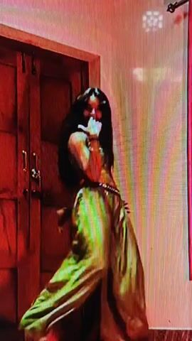A woman is dancing in front of a wooden door and a pink curtain. She has long, dark hair and is wearing a gold-colored top and loose green pants. Her dance moves are fluid and expressive, with a focus on her arms and hips. The lighting is dim, with a reddish tint. The scene appears to be indoors, possibly a home or a studio.