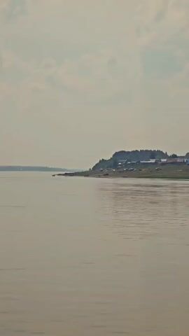The video shows a wide, calm river with a gentle ripple. On the far bank, a green, gently sloping hill is dotted with numerous small buildings, suggesting a village or settlement. Trees are visible in the background, and the sky is overcast with a few light clouds. The overall impression is one of a serene, rural landscape.