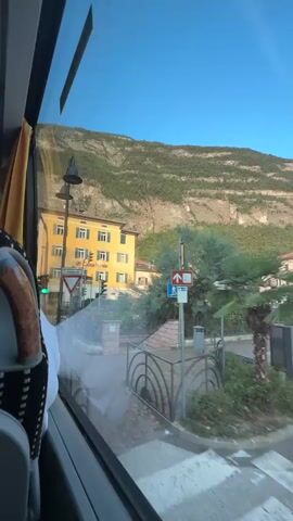 The video shows a view from the window of a vehicle, likely a bus. Outside, a yellow building with multiple floors and green shutters is visible, along with a traffic light that is currently green. A steep, tree-covered mountain rises behind the buildings. Several road signs are also visible, including a yield sign, a pedestrian crossing sign, and a sign with an upward arrow and a red circle with a diagonal line, indicating no entry for certain vehicles. The foreground shows part of the vehicle's interior, including a seat and the window frame. The lighting suggests it is daytime, possibly late afternoon or early morning, with clear blue skies.
