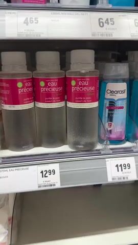 The video shows a retail store aisle. The camera pans across shelves stocked with various beauty and personal care products. In the foreground, on the first shelf, are several bottles of "EAU PRÉCIEUSE Lotion," priced at €12.99. To the right of these bottles is a bottle of "Clearasil ULTRA Rapid Action Lotion" with a price tag of €11.99. On the shelf below, there are containers of "DEMAK up SENSITIVE" makeup remover pads, priced at €4.29. Further down the aisle, more products are visible, but they are out of focus. The lighting appears to be standard fluorescent store lighting. There are no people visible in the video. The focus is entirely on the products displayed on the shelves.