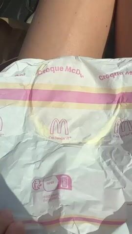 The video shows a person holding a McDonald's Croque McDo sandwich in its paper wrapper. The sandwich appears to be made with two toasted buns, ham, and melted cheese. Some of the cheese is oozing out from the sides of the sandwich. The wrapper has the McDonald's logo and the slogan "I'm lovin' it." The person is sitting in a car, with their legs visible in the lower part of the frame. There is also another McDonald's bag in the background. The lighting suggests it is daytime.