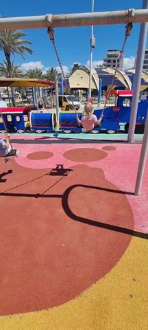 A young child, seen from behind, is swinging on a swing in a playground. The child has blond hair pulled back in a small ponytail and is wearing a light pink t-shirt and patterned leggings with green shoes. The swing has metal chains. Another person can be seen swinging on another swing to the left of the frame. The playground has a red and pink rubber surface with circular patterns. In the background, there is a blue and red train-themed structure, palm trees, and buildings under a clear blue sky. A yellow van is also visible in the background. The scene is brightly lit, with the shadows of the swing and supporting structures prominent on the ground.