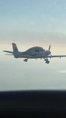 The video displays a single-engine aircraft in mid-flight, viewed from another aircraft. The scene is set against a clear sky with subtle cloud formations, suggesting it is daytime. The sun appears to be low on the horizon, possibly indicating sunrise or sunset, casting a warm glow. The aircraft, a white composite plane, has the registration number "N219EL" clearly visible on its fuselage. The camera appears to be capturing the footage through a window, with some smudges or reflections visible on the glass. The overall mood is serene and peaceful, capturing a moment of flight. The location is identified as Santa Barbara, United States of America.