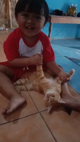 A child in a red and white shirt is sitting on the floor with an orange tabby cat. The child is holding the cat's front paws and gently pulling them. The cat appears relaxed and is looking at the child. The floor is tiled with a light brown color. In the background, there is a blue wall and some furniture. The overall scene depicts a playful interaction between a child and a pet.