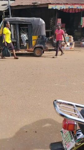 The video shows a street scene in a tropical environment. In the foreground is the rear of a parked motorcycle, with a chrome luggage rack and taillight visible. The road is paved, but appears rough and possibly made of red soil. Across the street, a small, open-fronted shop is packed with stacked goods, including boxes, bottled drinks, and hanging snacks. The shop's roof is corrugated metal with colorful advertising banners. Several people are walking on the street. One person is walking toward the shop, another is walking away from it. In the background, palm trees and other vegetation suggest a tropical climate. The lighting is bright and suggests a sunny day. A motorcycle is parked near the shop. A yellow autorickshaw is partially visible on the left.