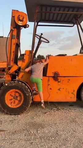 A small child with blonde hair and wearing a grey t-shirt, green shorts, and sneakers is climbing onto an orange forklift. The forklift has large black tires, a metal cage overhead, and a steering wheel visible inside. The child is using handholds and footrests on the side of the machine to ascend. The setting appears to be an outdoor gravel area with a building and some lumber visible in the background under a partly cloudy sky.