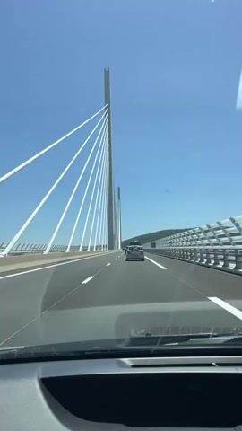 The video shows a first-person view from inside a car traveling on a highway that is on a cable-stayed bridge. The sky is clear and blue, and the sun is shining. White cables descend from tall, slender towers to support the bridge deck. Other vehicles are visible on the highway ahead, including a silver SUV. The dashboard of the car is visible in the foreground, showing a digital display that reads "85". The road has multiple lanes separated by white lines. To the right of the highway, there is a metal barrier and further to the right, a hillside covered with trees. The bridge appears to be very high above the ground, given the height of the towers and the distant view of the landscape.