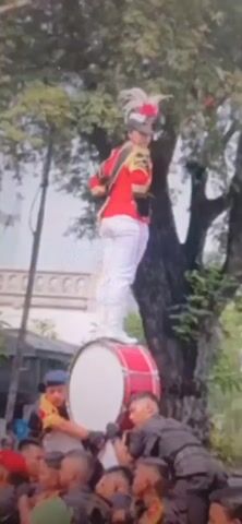 A person in a red and gold uniform with a feathered hat stands on top of a large drum. The person is facing away from the camera, looking towards a tree. Below the drum, two people in dark uniforms are holding the drum steady. The background shows a building and more trees.