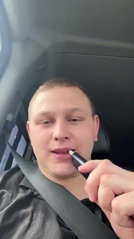 The media file appears to be a first-person perspective video captured from inside a vehicle, likely a car or truck. The initial shot focuses on a person from the chest up, wearing a black collared shirt and a seatbelt. The person is holding a dark-colored cylindrical object to their lips, suggesting they might be vaping or smoking. Their gaze is directed upwards and to the right, with a neutral expression. A partial view of a tattoo is visible on their neck.

The subsequent frames shift to outward views from the vehicle's window. The first exterior shot shows the side of a large truck passing by, identified by a red and black design and a partial registration plate that reads "CZ". The second exterior shot reveals a rural landscape with fields of dry grass or wheat, a line of trees in the distance, and a hazy, overcast sky, suggesting it might be daytime with cloudy weather. The final exterior shot shows the side of a large, grey trailer truck, again with a visible tire and wheel assembly. The overall impression is of travel on a highway in a rural area, possibly in the Czech Republic, given the "CZ" license plate fragment. The time of day is difficult to ascertain precisely due to the overcast conditions but appears to be daylight.