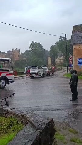 A bus is driving on a wet road. There is a car being towed behind a truck. Two people are standing on the side of the road watching. There is a bicycle in the foreground.