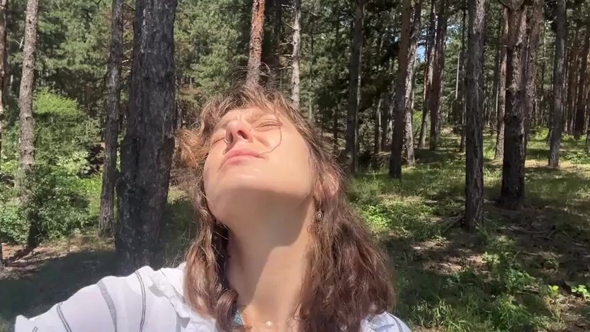 A medium shot shows a young woman with light brown hair and a white shirt standing in a forest on a sunny day. She is looking upwards with her eyes closed and a slight smile on her face, seemingly enjoying the sun. Her hair is slightly windswept, and she wears a delicate pearl necklace and earrings. The background is filled with numerous tall pine trees and green undergrowth, bathed in bright sunlight, creating a serene and natural setting.