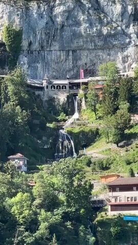 The video shows a wide shot of a cliff face with a building integrated into the rock. Waterfalls cascade down the cliff face in front of the building. There are trees and some houses visible on the slopes below the cliff. The lighting suggests it is daytime with bright sunlight. There are no people or animals visible in the frame. The scene appears to be a natural landscape with some human architectural elements.