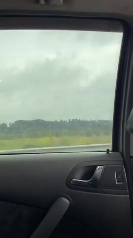 The video shows the interior of a car, specifically the rear passenger door and window, looking out at a landscape. The car is in motion, as indicated by the blurred road and passing scenery. Outside, a green, hilly landscape with trees and a cloudy sky is visible. The car's interior features a dark gray door panel with a chrome door handle, a window control button, and a fabric pattern on the lower section. A faint reflection of the car's interior can be seen on the window. The overall weather appears to be overcast or rainy.