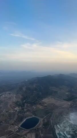 This is an aerial video taken from a hot air balloon above the countryside near Aspe, Spain. The scene is a vast, mountainous landscape bathed in the soft light of early morning or late evening. The dominant subjects are the rugged, dark green mountains that stretch across the horizon, appearing layered and distant due to atmospheric haze. In the foreground, the terrain is a mosaic of dry, cultivated fields and patches of vegetation, crisscrossed by dirt roads and dotted with scattered buildings and agricultural structures.

A prominent circular reservoir or pond, dark blue in color, is a key focal point in the middle ground, surrounded by fields and infrastructure. The landscape shows signs of human activity, including what appears to be a quarry or industrial site on the right side of the frame, with lighter colored earth and machinery visible.

The sky above is a gradient of soft blues, with streaks of pale yellow and white clouds indicating either sunrise or sunset. The overall atmosphere is serene and expansive, conveying a sense of natural beauty and the quietude of rural life. There is no visible text in the video. The time of day is suggested by the low angle of the sun and the soft, diffused light, creating long shadows and a muted color palette. The weather appears clear, with the haze suggesting a dry climate common to the region.