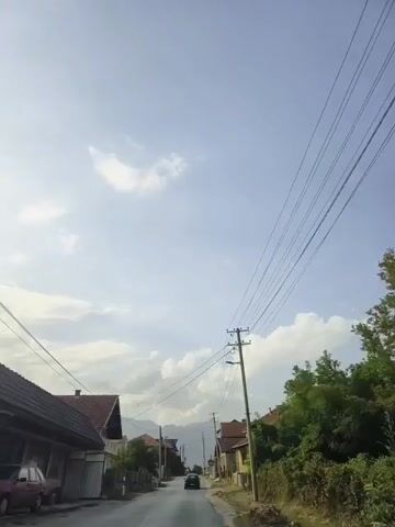 This is a first-person perspective video captured while driving down a rural road lined with houses and trees. Overhead, a web of power lines crisscrosses the sky, supported by utility poles. A single car is visible in the distance on the road. The sky is partly cloudy.