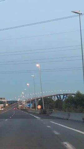 The video shows a first-person perspective of driving on a highway at dusk. The road is paved and marked with white lines. There are streetlights lining the highway, and they are illuminated. In the distance, a large bridge with lights is visible. There are a few cars on the highway, and they appear to be driving at a moderate speed. The sky is a pale blue, indicating that the sun has recently set. There is a speed limit sign that reads 90. There are also directional signs visible. The overall impression is of a peaceful drive on a highway as evening approaches.