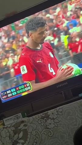 A man in a red soccer jersey with the number 4 on the back stands on a field holding a soccer ball. He has dark curly hair and a mustache. He is looking down at the ball. A scoreboard in the lower left corner of the screen shows the score of a soccer game between Australia (AUS) and Egypt (EGY). The score is AUS 2, EGY 3. The man is holding the ball with his right hand and appears to be adjusting it. The background shows a crowd of people in a stadium.