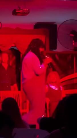 The video shows a drag performance taking place on a stage lit with red lights. In the first frame, a performer with long dark hair is wearing a light blue top and a sparkly dress. They are holding a microphone and appear to be singing or speaking. The stage has red curtains in the background and a large fan is visible on the right side of the stage. The second frame shows the same performer, with their head turned and their hands raised. The third frame shows a performer in a dark shirt on stage, with their arms raised, and the words "GRAND SHOW" are visible above them. The fourth frame shows a performer sitting in a chair on stage, wearing a yellow shirt and a red overall dress with dark pigtails. They are looking to the side. The fifth frame shows a performer wearing a blonde wig and a dark dress, with their hand raised to their face. The overall scene suggests a lively performance in a dimly lit venue, likely a club or theater.