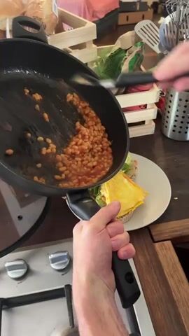 A pair of hands is shown tilting a frying pan over a plate of food. The frying pan contains baked beans in a tomato sauce. The plate holds two open-faced sandwiches topped with lettuce, avocado, and a folded omelet. One hand grips the handle of the frying pan, while the other hand uses a spatula to help guide the beans onto the sandwiches. The background includes a kitchen counter with a wooden crate filled with produce and kitchen utensils in a holder. The scene appears to be a home kitchen.