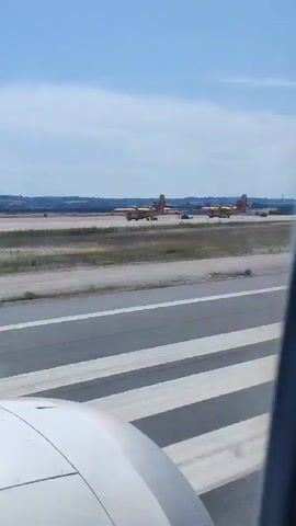 This video shows a view from an airplane window, looking out at an airport and then the coastline. At the beginning of the video, several yellow aircraft, possibly Canadair CL-415 firefighting planes, are visible on the tarmac. The plane from which the video is filmed is on the runway, and the wing or engine cowling is visible in the foreground. The scene then shifts to show a wide expanse of ocean and a coastal town under a bright, partly cloudy sky, suggesting it is daytime. The weather appears to be clear and sunny. The camera pans across the coastline, revealing buildings and a beach area. No text is visible in the video. The overall impression is one of departure or arrival at an airport located near a coastal area, likely Thermi, Greece, given the context. The video captures a transition from an airport environment to a scenic coastal view.